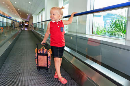 Happy little child with luggage stand on airport transit hall walkway moving to plane departure gate for waiting flight boarding. Active family lifestyle, travel by air with kid on summer holiday tourの写真素材