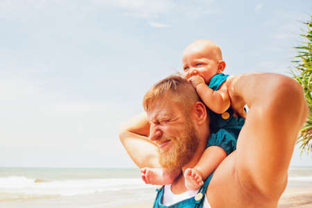 Funny face portrait of happy family - baby son sit on father shoulders, walk with fun along sea surf on sunny beach. Active parents, people activity on summer vacation with child on tropical island.の写真素材