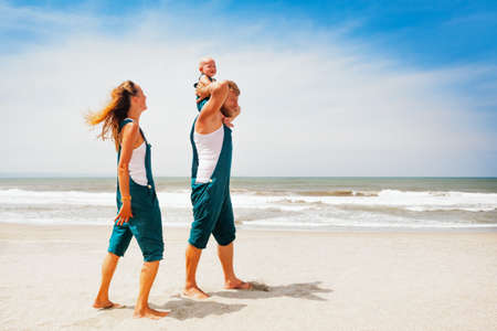 Funny portrait of happy family. People walk with fun along sea surf on sunny beach. baby son sit on father shoulders, look at mother. Leisure activity on summer vacation with child on tropical island.の写真素材