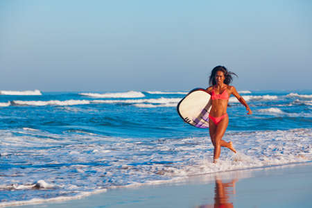 Girl in bikini with surf board has fun. Woman surfer run into water, jump with splashes through ocean wave. People in water sport adventure camp, beach extreme activity on summer beach family vacationの写真素材