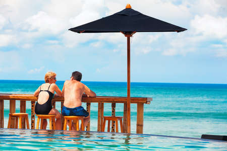 Successful retirement recreation, summer vacation concept. Retired mature couple enjoying beautiful sunny day in swimming pool at beach club. Happy senior woman and man sitting at poolside beach bar.の写真素材