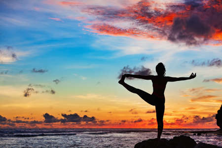 Sunset meditation silhouette. Active woman stand in yoga pose on beach rock to keep fit and health. Healthy lifestyle, fitness training, sport activity on summer family holiday.の写真素材