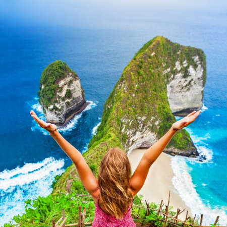 Family vacation lifestyle. Happy woman with raised in air hand stand at viewpoint. Look at beautiful beach under high cliff. Travel destination in Bali. Popular place to visit on Nusa Penida islandの写真素材
