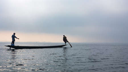 Black silhouette of two fishers standing on stern and paddling by legs, travelling by traditional...
