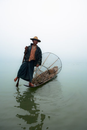 Inle Lake, Myanmar - January 04, 2007: Local fisher with traditional Burmese fishing net standing on stern of long boat and rowing oar by leg, on Inle lake, Myanmar.のeditorial素材