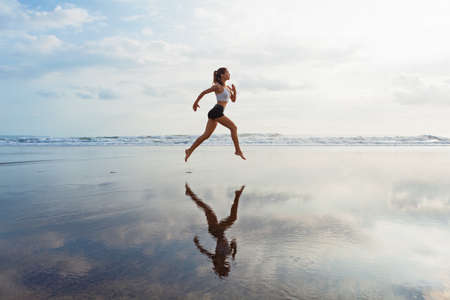 Barefoot sporty girl with slim body running along sea surf by water pool to keep fit and health. Beach background with blue sky. Woman fitness, jogging sports activity on summer family vacation.の写真素材