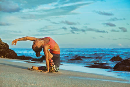 Meditation on sunset sky background. Young active woman sit in yoga pose on sand beach, stretching to keep fit and health. Healthy lifestyle, outdoor fitness, sports activity on summer family holiday.の写真素材
