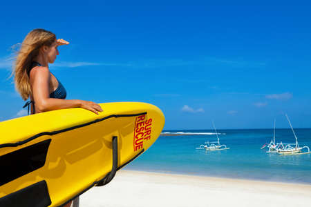 Life saving yellow board with surf rescue sign. Young lifeguard woman stand on duty, look at blue...