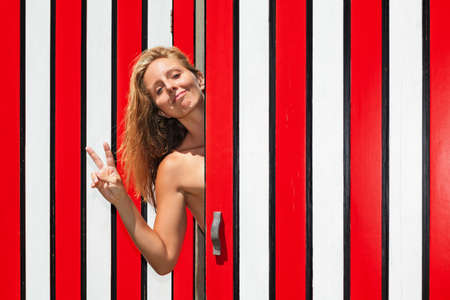 Happy smiling young blond woman against red striped wall of beach changing cabin has fun, raising hand and showing v-sign victory or peace with fingers gesture. Family summer holiday. Positive people.の写真素材