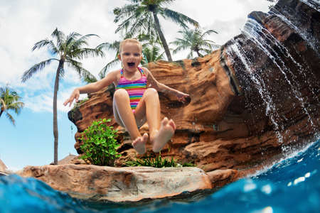 Funny child learn to swim with fun. Girl jumping high with splashes into water pool under waterfall....