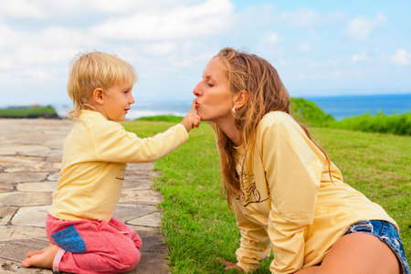 Happy family on outside walk. Child sitting on green grass lawn have fun. Young attractive mother kiss son finger. Positive people activity on tropical vacation with kids. Summer lifestyle backgroundの写真素材