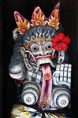 Old figure of traditional Balinese hindu spirit Rangda on wall of village temple at Bali island. Decorated by red flower for religious festival at Galungan holiday. Art, culture of Indonesian people.の写真素材