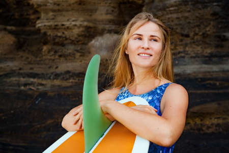 Sporty girl in bikini with surfboard stand by black cliff on beach. Surfer woman look at sea surf...