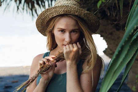 Young beautiful hipster woman in straw hat hold in hands bouquet of dried wild flowers. Happy romantic girl walk on tropical sea beach. Travel lifestyle, summer vacation accessories trend - boho styleの写真素材