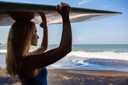 Girl in bikini with surfboard walk on black sand beach. Surfer woman look at sea surf and water pool with white foam. Active people in sport adventure camp, extreme activity on summer family vacationの写真素材