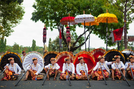 Bali, Indonesia - June 23, 2018: Group of young dancer men in ethnic costumes with traditional red, black and yellow umbrellas on hindu ceremony parade during temple festival. Balinese people culture.のeditorial素材