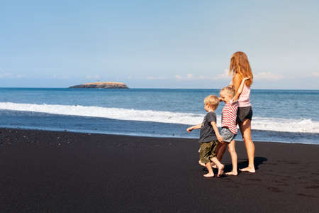 Happy family - young mother, daughter, baby son hold hands, walk together by water pool along sea surf on black sand beach. Travel, active lifestyle, parents with child on summer vacationの写真素材