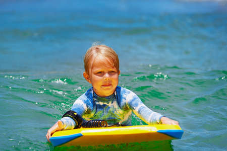 Happy baby girl - young surfer ride on surfboard with fun on sea waves. Active family lifestyle,...