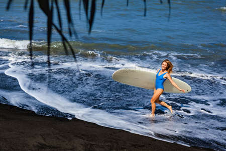 Sporty girl in bikini with surf board walk on black sand beach. Young surfer woman run by water pool...