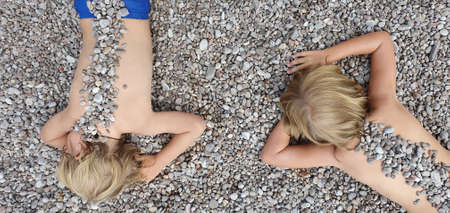 On sunny beach happy kids have fun, lying down on hot pebble, sunbathing, warming after swimming in sea. Active children lifestyle, summer vacation travel with kids on tropical family resort.の写真素材