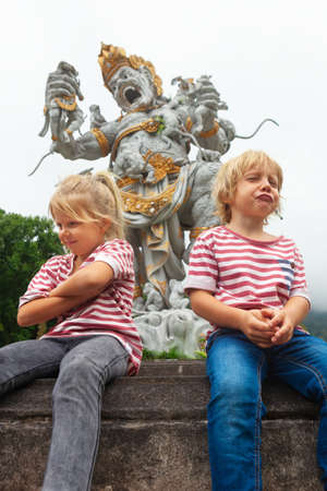 Funny little kids in botanical garden on background of ancient statue of fighting Kumbakarna from epic Hindu legend Ramayana. Traditional arts, culture of Bali, popular travel destination in Indonesiaの写真素材