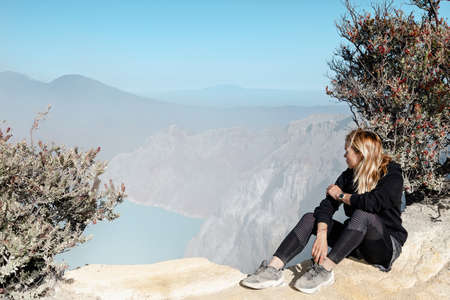 Happy young woman sit on rock above volcano Kawah Ijen crater. Look at largest in world acidic lake,...