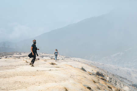 Young women in protective mask walking by wastelands around Kawah Ijen volcano crater. Post apocalypse landscape with clouds of toxic gases from volcanic emissions and dead land poisoned by sulphur.の写真素材
