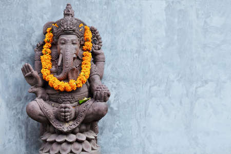 Ganesha sitting in meditating yoga pose in front of hindu temple. Decorated for religious festival by orange flowers garland, ceremonial offering. Balinese travel background. Bali island art, culture.の写真素材