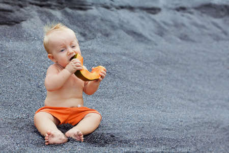 Funny photo of positive baby boy with smiling face eating ripe fruit - orange papaya slice. Healthful food, natural breakfast on sea black beach. Healthy lifestyle on summer family holiday with kids.の写真素材