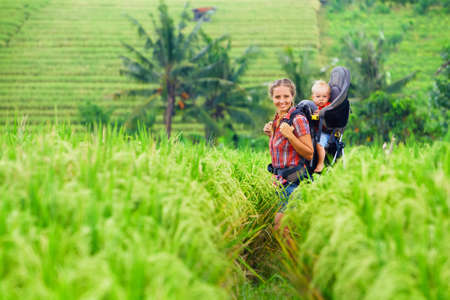 Nature walk in green rice terrace field. Happy mother hold little traveller in carrying backpack. Baby ride on woman back. Travel adventure, hiking with child carrier, family summer vacation on Bali.の写真素材