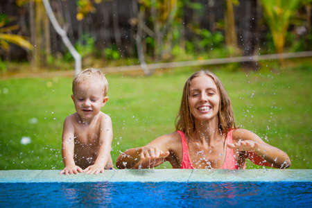 Family funny holiday. Happy son with mother have fun in swimming pool in tropical garden. Active baby splashing at poolside. Summer healthy lifestyle, kids water activity, games, lessons with parents.の写真素材
