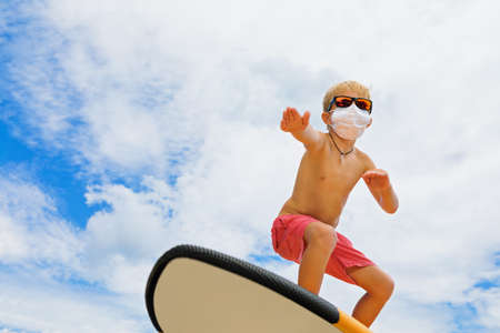 Young funny surfer wearing sunglasses, protective face mask ride on surfboard. Summer beach tours, cruises cancelled due to coronavirus COVID-19 epidemic. Safe travel destinations for family vacation.の写真素材