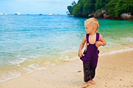 Happy barefoot child in loose jumpsuit have fun on tropical sea beach. Funny baby boy run by water along surf edge. Active kids lifestyle, people activity on family summer holiday with childrenの写真素材