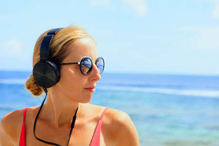 Young positive girl in headphones and sunglasses listening music and relaxing. Woman chilling at tropical sea beach. Travel family lifestyle, recreational activities at summer cruise vacations.の写真素材