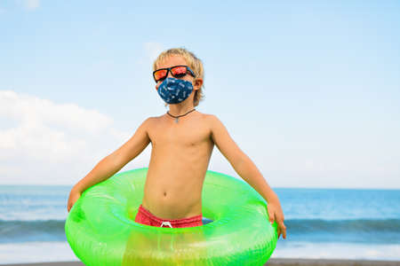 Funny child in sunglasses, inflatable ring on tropical sea beach. New rules to wear cloth face covering mask at public places due coronavirus COVID 19の写真素材