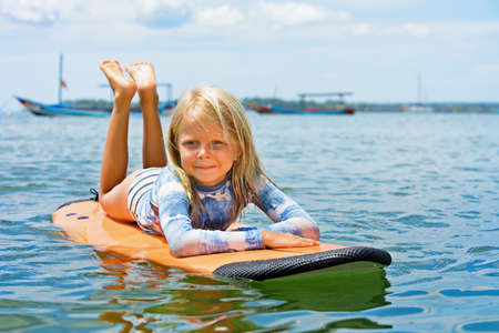 Happy baby girl - young surfer learn to ride on surfboard with fun on sea waves. Active family lifestyle, kids outdoor water sport lessons, swimming activity in surf camp. Summer vacation with child.の写真素材