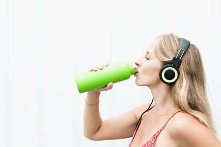 Young blond woman on white background drink fresh spring water from green reusable bottle. Healthy...