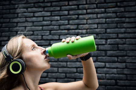 Young blond woman on black background drink fresh spring water from green reusable bottle. Healthy lifestyle in summer city.の写真素材
