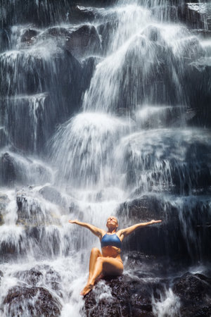 Travel in Bali jungle. Beautiful young woman sitting on rock under falling spring water, enjoy tropic cascade waterfall. Asian nature, day trip, walking adventure, fun on family summer vacationの写真素材