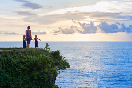 Happy children with mother have fun on summer beach holiday. Funny little kids stand at sea cliff. Looking at beautiful view of sunset sky. Healthy family lifestyle, summer travel on tropical island.の写真素材