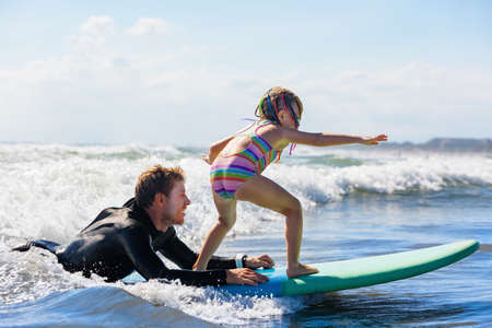 Little surf girl - young surfer learn to ride on surfboard with instructor at surfing school. Active...