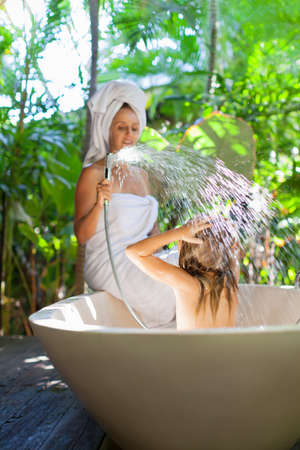 Happy baby girl with mother have fun in bath. Playful woman spraying child from shower in outside bathroom on open veranda with beautiful tropical garden view in luxury villa on summer family vacationの写真素材