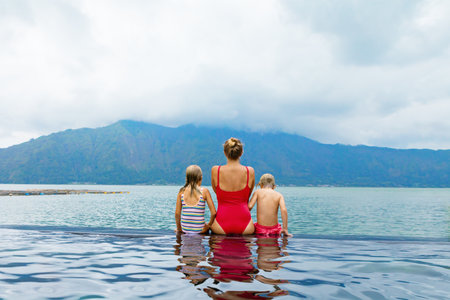 Happy family in Batur volcano hot spring spa. Travel in Kintamani, Bali. Mother, kids chilling in infinity pool with lake view. Healthy lifestyle, recreational activity on summer holiday with childrenの写真素材