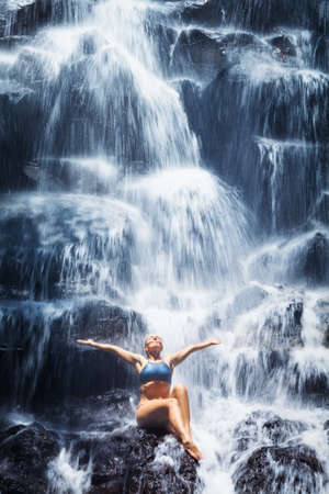 Travel in Bali jungle. Beautiful young woman sitting on rock under falling spring water, enjoy tropic cascade waterfall. Asian nature, day trip, walking adventure, fun on family summer vacationの写真素材