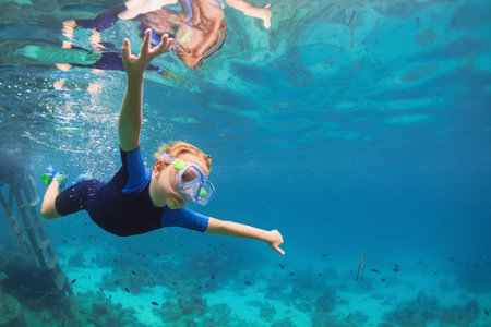 Happy family - active kid in wetsuit and snorkeling mask dive underwater, see tropical fishes in coral reef sea pool. Travel adventure, swimming activity on summer beach vacation with child.の写真素材