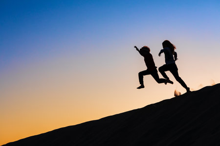 Black silhouette at sunset sky background. Happy family - kids running and jumping high in the air from sea beach sand dune. Active people, outdoor activity on tropical summer vacations with childrenの写真素材