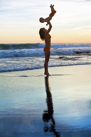 mother tossing up baby son into mid air, catching on sunset sky, sun backgroundの写真素材