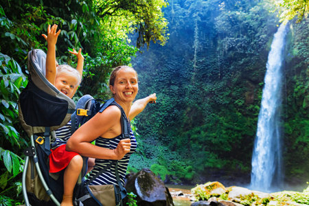 In waterfall pool young happy mother hold little traveller on back - baby girl in carrying backpack enjoying travel adventure, Hiking activity with child on family summer vacation, weekend nature tourの写真素材