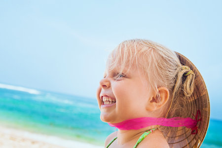 On sunny white sand beach happy smiling baby girl with vietnamese straw hat on head has fun before swimming in sea waves. Active travel family lifestyle, water activity on summer vacation with child.の写真素材
