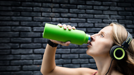Young blond woman on black background drink fresh spring water from green reusable bottle. Healthy lifestyle in summer city.の写真素材